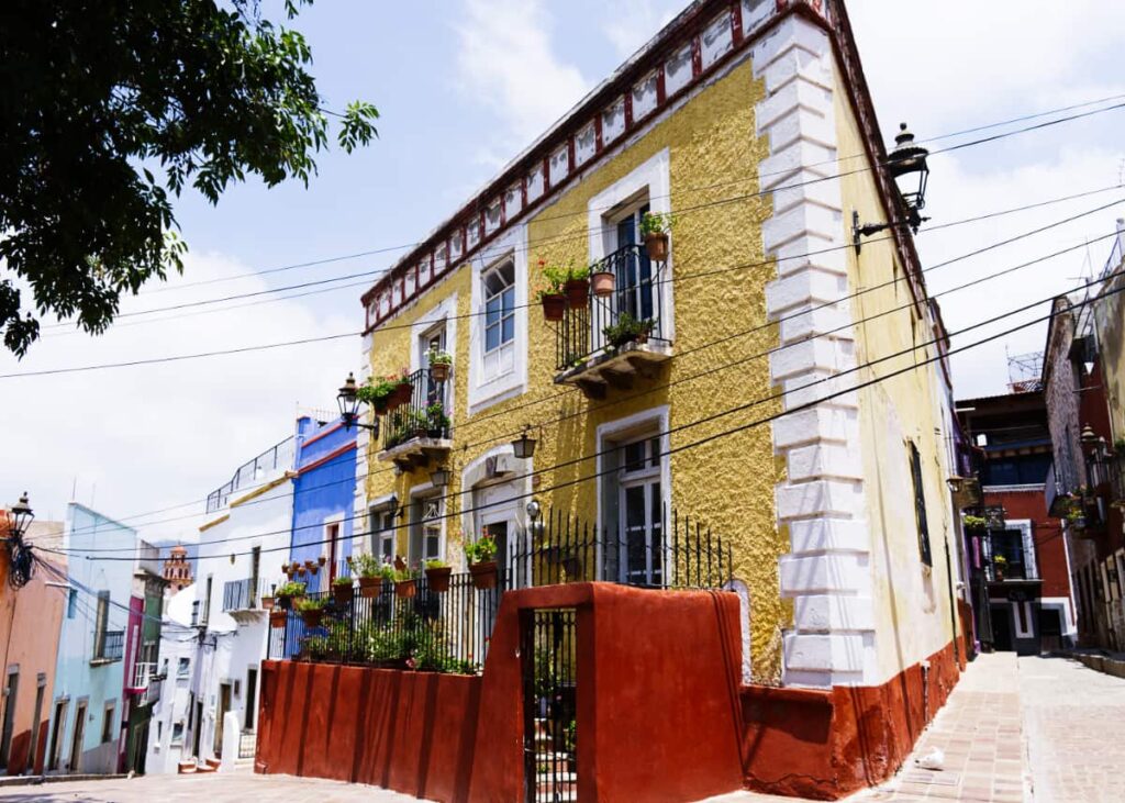Walking the alleys is one of the best things to do in Guanajuato City to see street scenes like this one. A bright yellow triangular house sits on the corner between two alleyways. The house is pained in red trim with white accents.