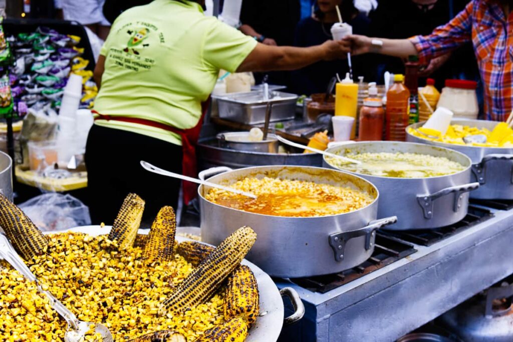 Large pots of corn, some grilled and some in various sauces line this Guanajuato street food stand. In the back ground, a woman hands a cup of elotes (a cup of corn kernals in broth) to a customer.