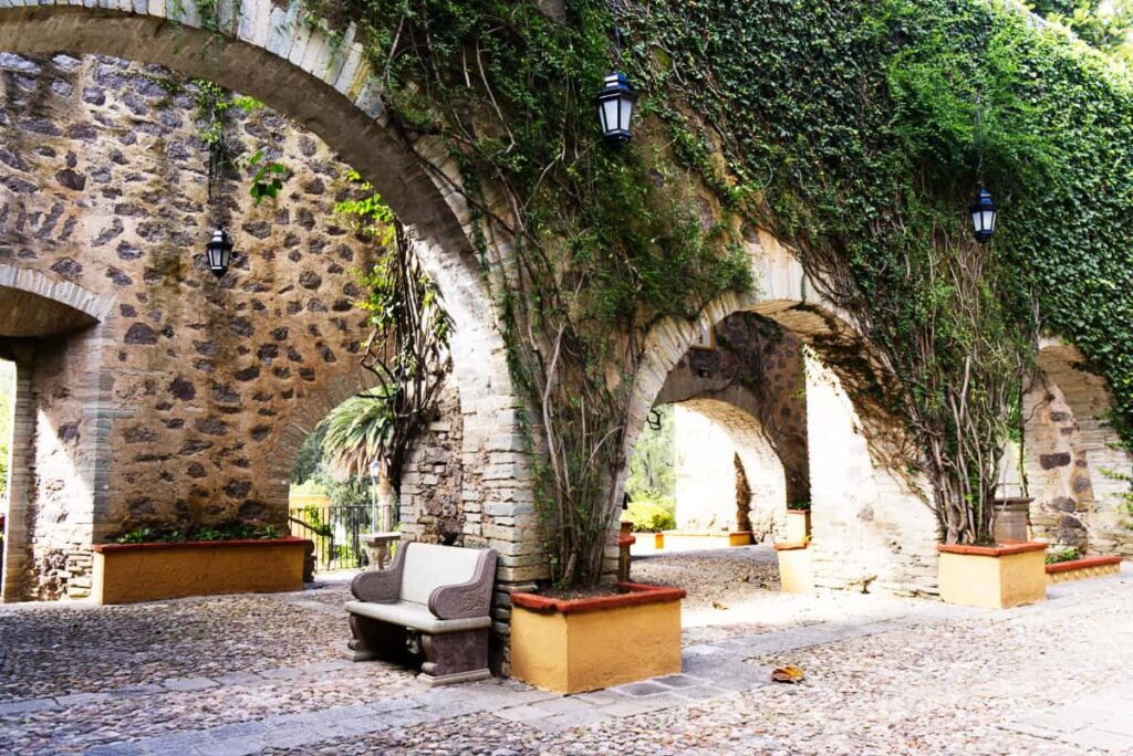 A peaceful scene at Hacienda San Gabriel de Barrera in Guanajuato capital features stone arches covered in climbing vines with lanterns and a stone bench.