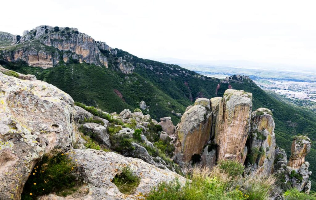 Views from the top of Cero La Bufa in Guanajuato show a large rocky mountain covered in green in the background and a cylindrical rock formation in the foreground. In the distance is a view of southern Guanajuato City.