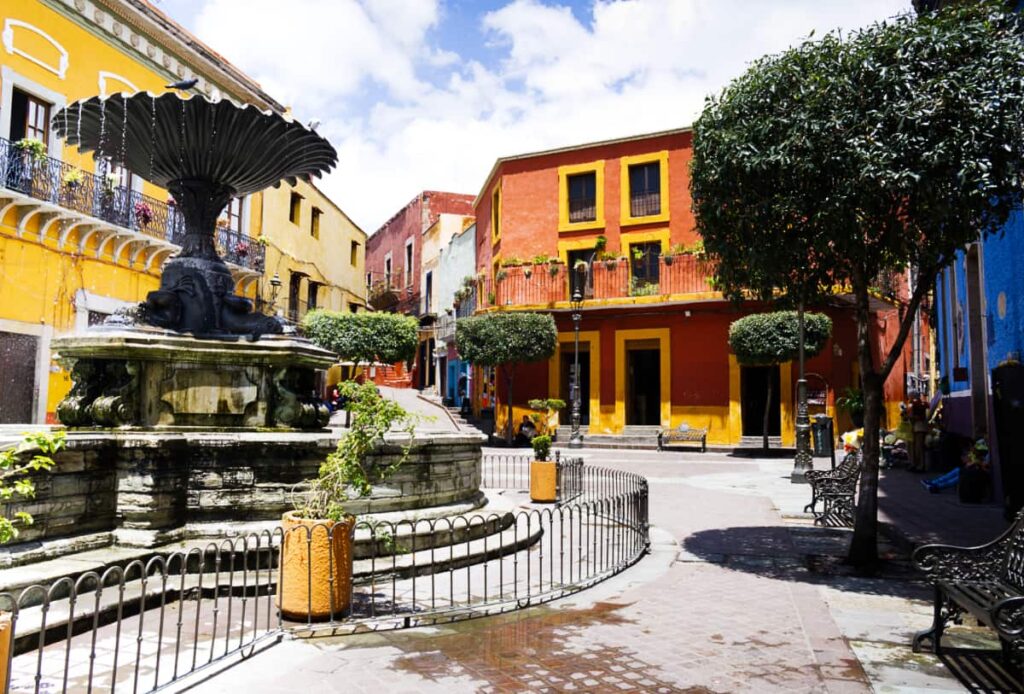 Guanajuato's Plaza Bartillo showcases a large iron fountain with three fish at the base. It's surrounded by colorful buildings, several iron benches, and callejones that lead in different directions.