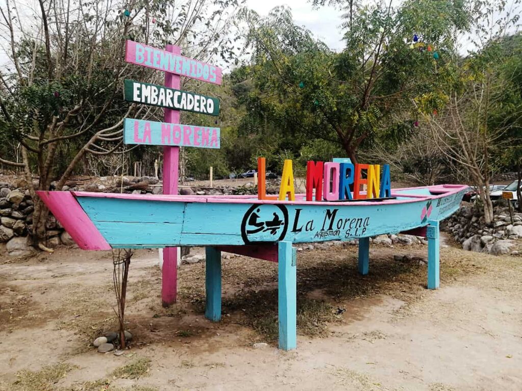 At La Morena, Tamul boat launch, a wooden turquoise boat with pink trim is set up on stilts. On top, multi color wooden letters read La Morena. A wooden beam behind has three signs that say "welcome," "boat launch," and "La Morena," in Spanish.