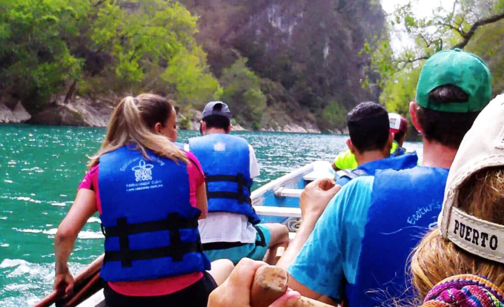 To reach the Tamul Waterfall, a group paddles along the Tampaon River. Everyone is wearing a blue life jacket with their backs facing the camera. In the background is a cliff face and the dark turquoise water.