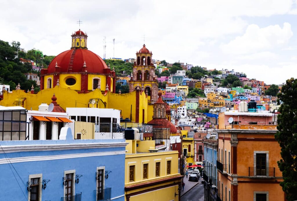Colorful buildings are built into the mountain in Guanajuato capital with a bright yellow and red dome of a church in the foreground.
