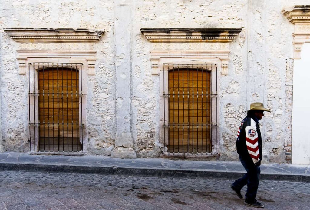 On the streets of Bernal Queretaro, a man wearing a cowboy hat and jacket walks in front of an old building with geometric details in the trim around the windows.