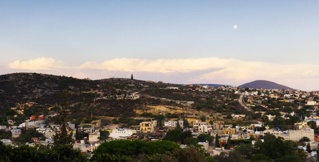 A full moon rises over Bernal Queretaro in the early evening. In the foreground are a hilly landscape and various buildings.