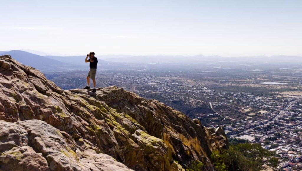 After climbing Pena de Bernal, a man uses binoculars to get a closer look. He is standing on a rocky ledge and behind him is a view of valley surrounding the pueblo magico.