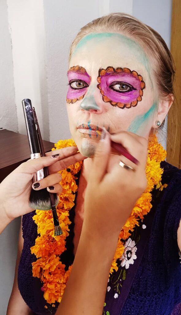 An artists paints a woman's face during a Oaxaca Day of the Dead tour. Her face has a slight covering of turquoise, her eyes are painted as pink and orange flowers. Around her neck is a necklace made of fresh marigold flowers.