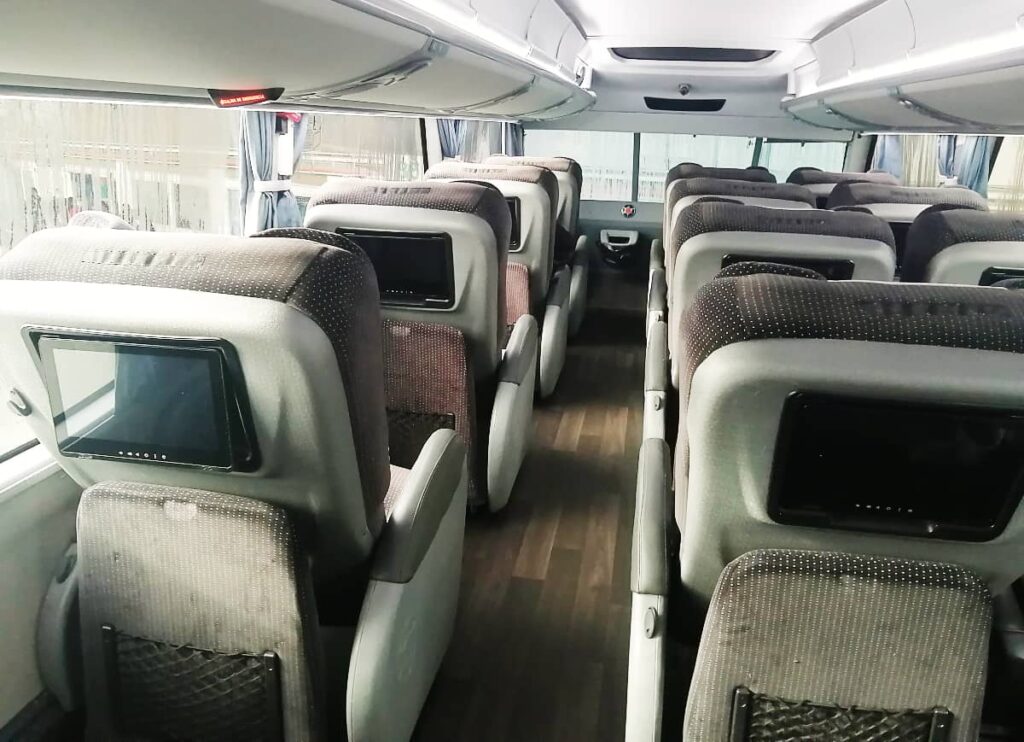A backview of the seats of the ETN luxury bus in Mexico. Each seat includes a foot rest and individual TV screen. The floor is wooden and there are overhead bins above the seats.