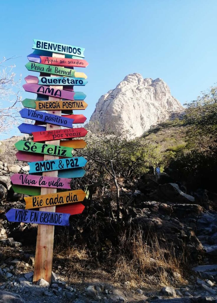 A post stands in front of Pena de Bernal Queretaro with colorful signs that say different phases in Spanish, such as Welcome, Positive Energy, Love and Peace, Positive Vibes.
