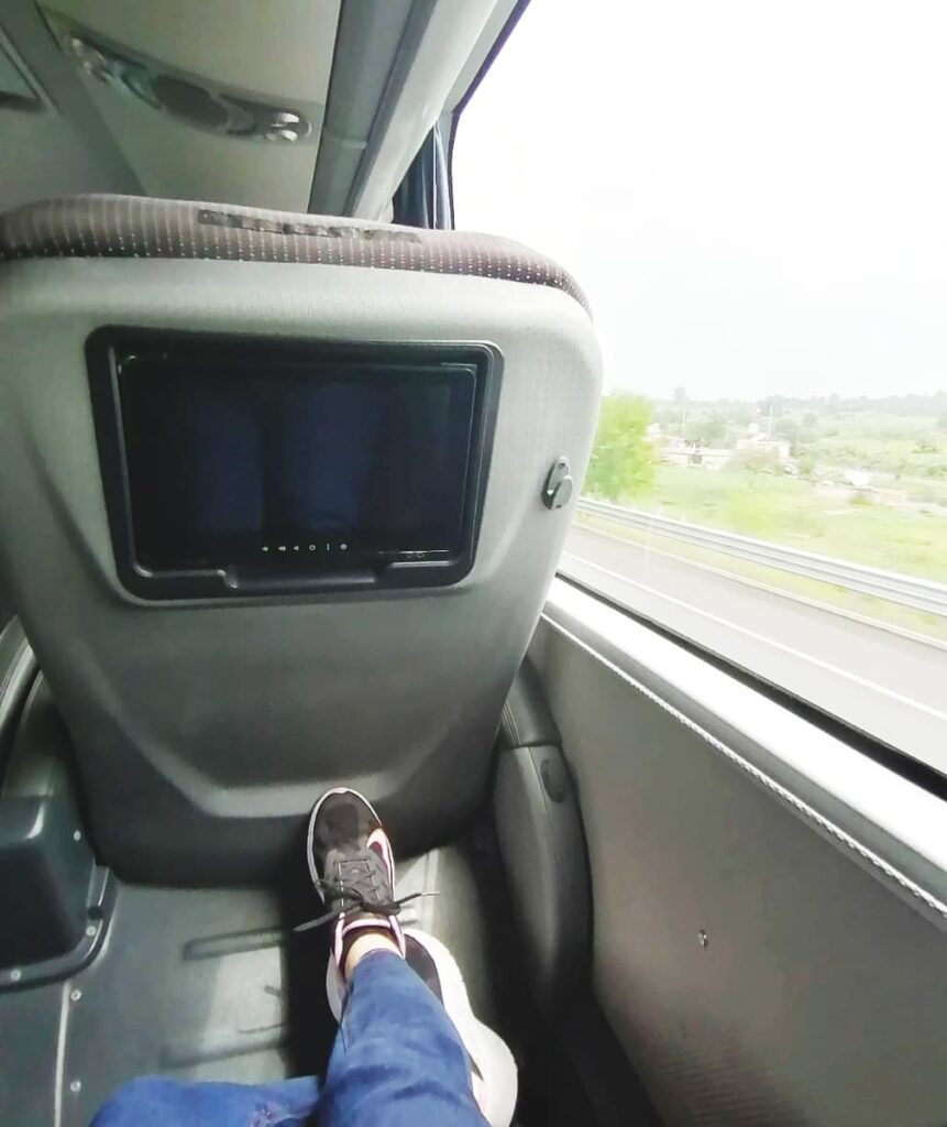 It is quite comfortable to travel Mexico by bus. This image shows two feet stretched out on the foot rest with an individual TV screen on the back of the chair in front. The large window gives the traveler views to the outside.