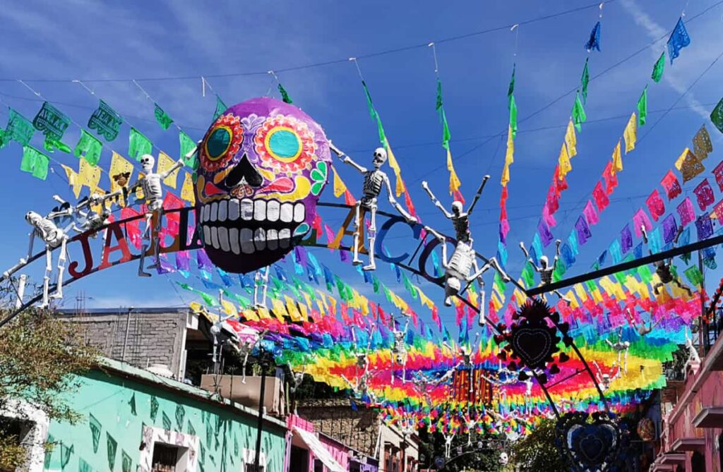 The arched sign of Jalatlaco, a place to stay in Oaxaca for Day of the Dead is decorated with colorful flags, small skeleton figures and a large catrina head.