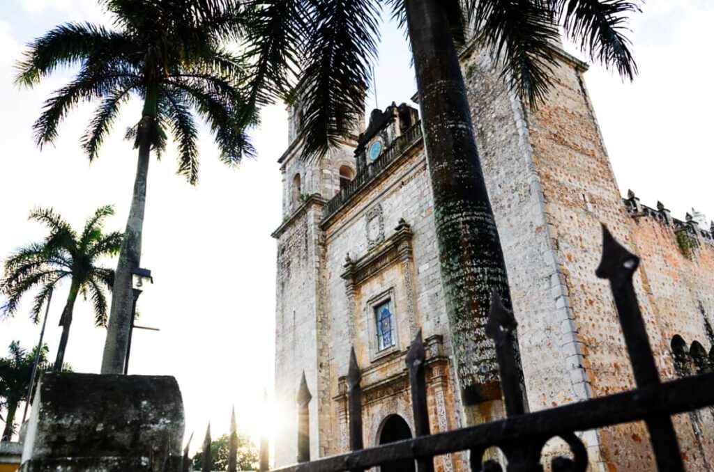 An upclose view of a stone church in Valladolid, Mexico surrounded by several tall palm trees. In the foreground is the black iron fence, with the sun shining just past the church.