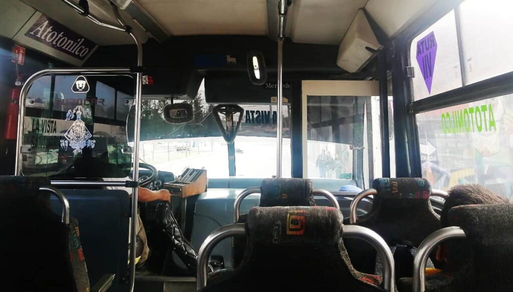 An inside view of the bus to La Gruta Spa from San Miguel de Allende. Seats are empty, with the driver's hand on the gear shifter and the words Atotonilco along both sides.