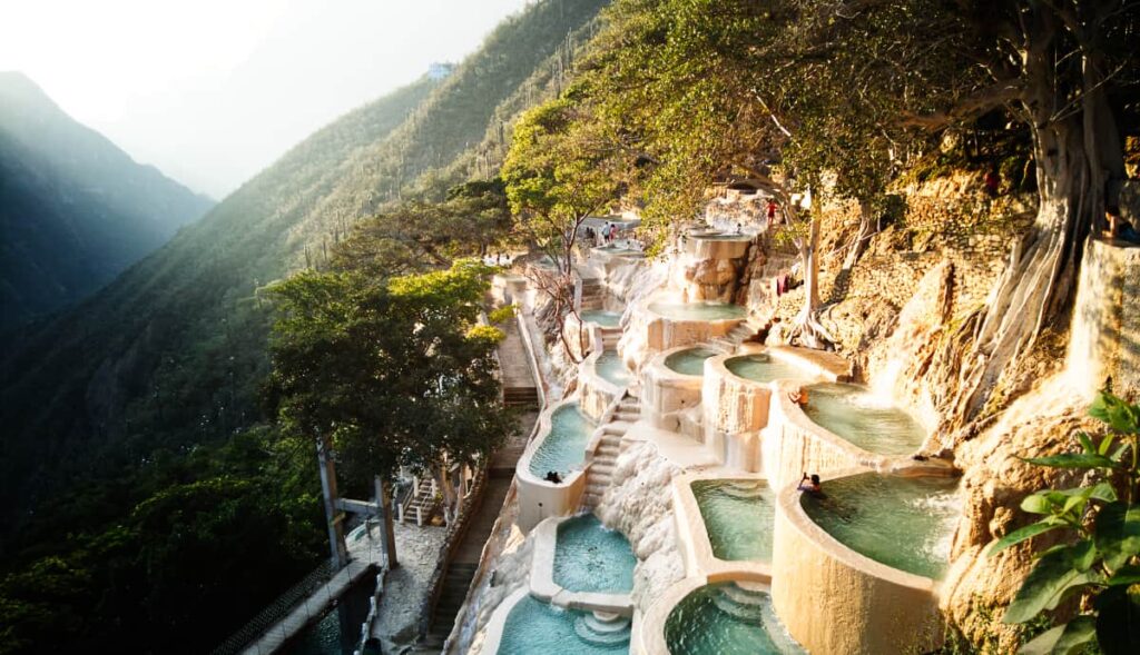 At Grutas Tolantongo, a series of manmade pools are layered on the side of the mountain mixed with several trees. The early morning light illuminates them as a couple of people swim.