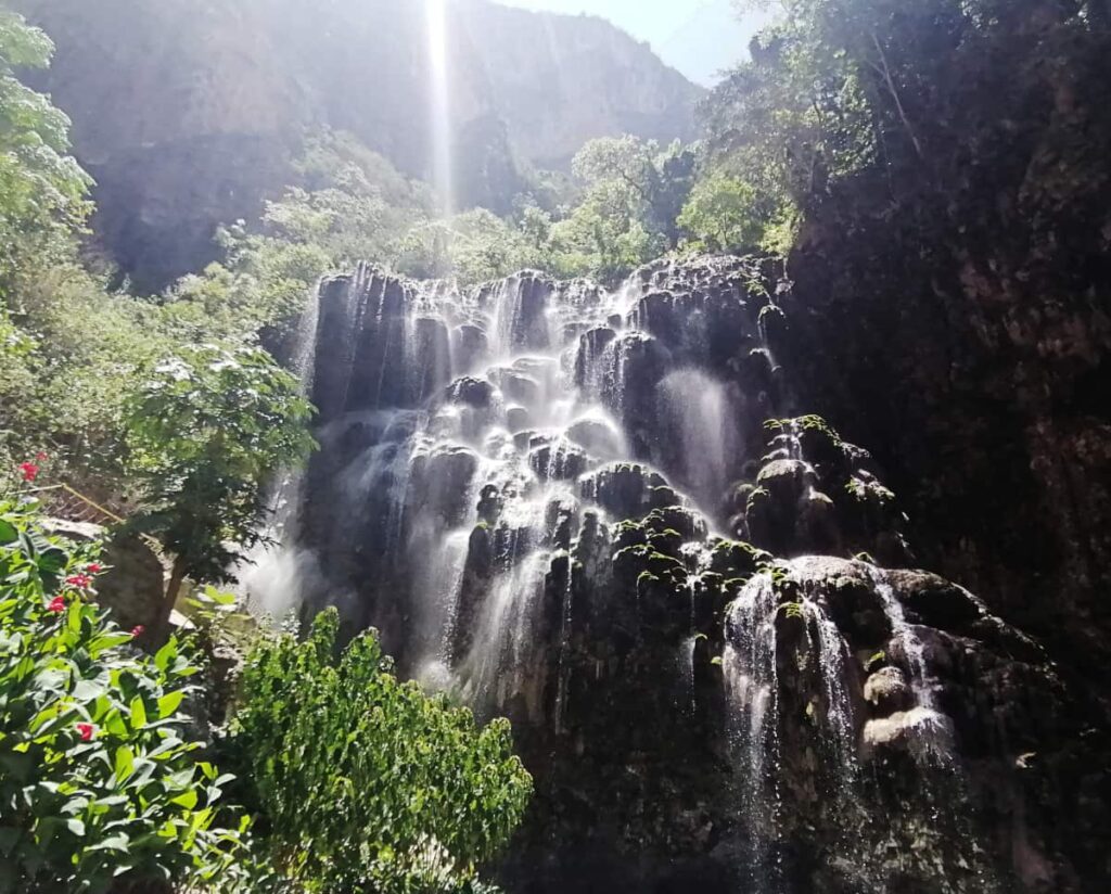 At the cave of Grutas Tolantongo, various streams of water fall down the rockface from the top to create a misty waterfall. The sun streams into the camera lens at the edge of the frame.