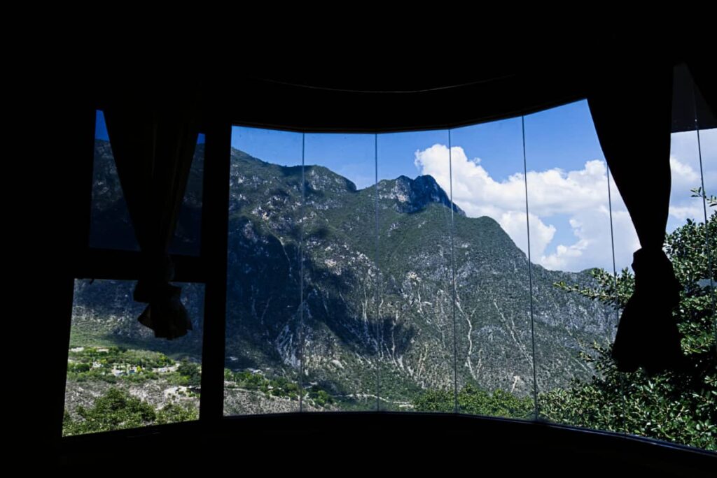 The curved window at a Grutas Tolantongo hotel room reveals a rugged mountain view with clear blue skies and white clouds