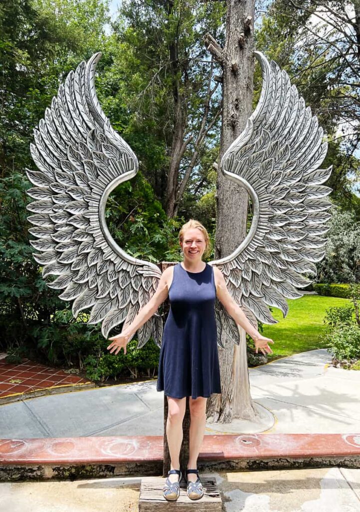 At La Gruta San Miguel de Allende, a blonde woman spreads her arms in front of a metal pair of angel wings. In the background are several trees.