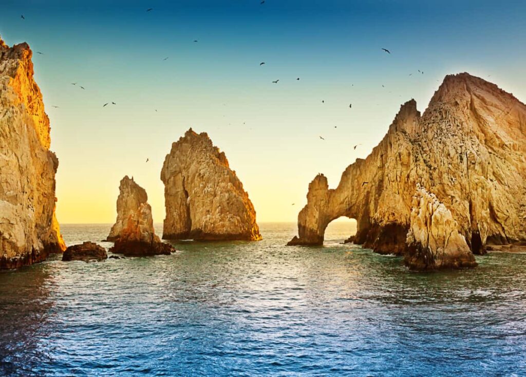 El Archo, a natural rock arch that makes Cabo one of the most beautiful cities in Mexico, is bathed in golden sunlight. Below is calm ocean waters and a dozen birds fly in the sky above.