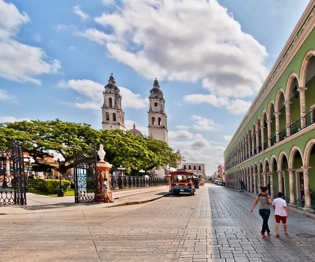 In Campeche, one of the pretties cities in Mexico, a green building with more than a dozen arches on both the first and second level line the street. Across, in the plaza are trees and a colonial church.