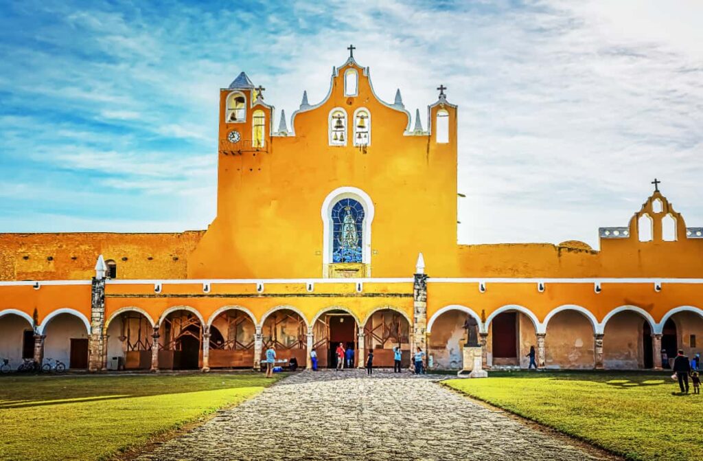People walk in front of a bright yellow church that features at least a dozen arches below in the yellow city of Izamal, one of the most beautiful towns in Mexico.