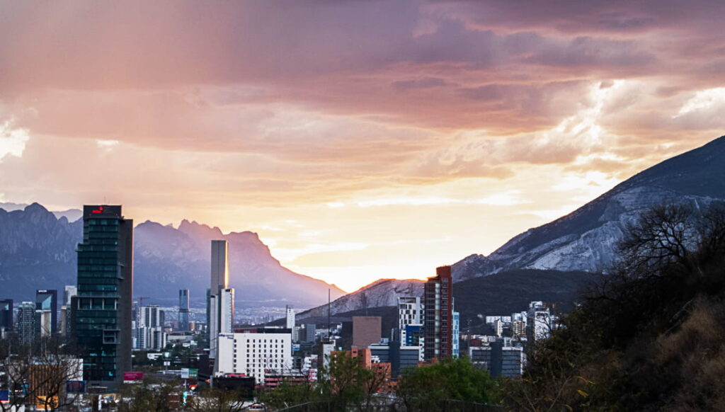 In the modern Mexican City of Monterrey, tall skyscrapers are contrasted with rugged mountains under a stormy sunset sky.