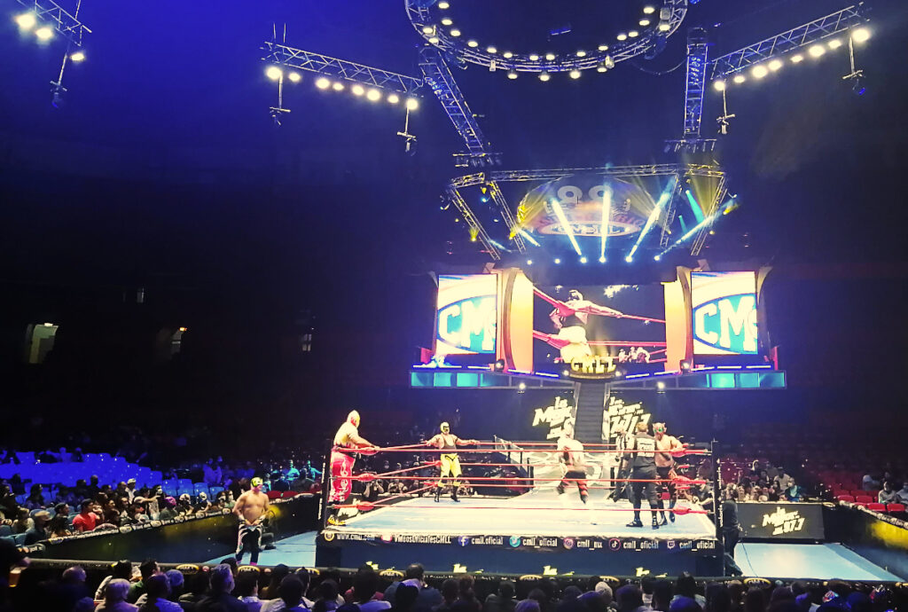 At Arena Mexico, wrestlers and a referee stand in the corners and middle of the stage at lucha libre in Mexico City. Behind them are large screens and lights. Around the stage are seated audience members.