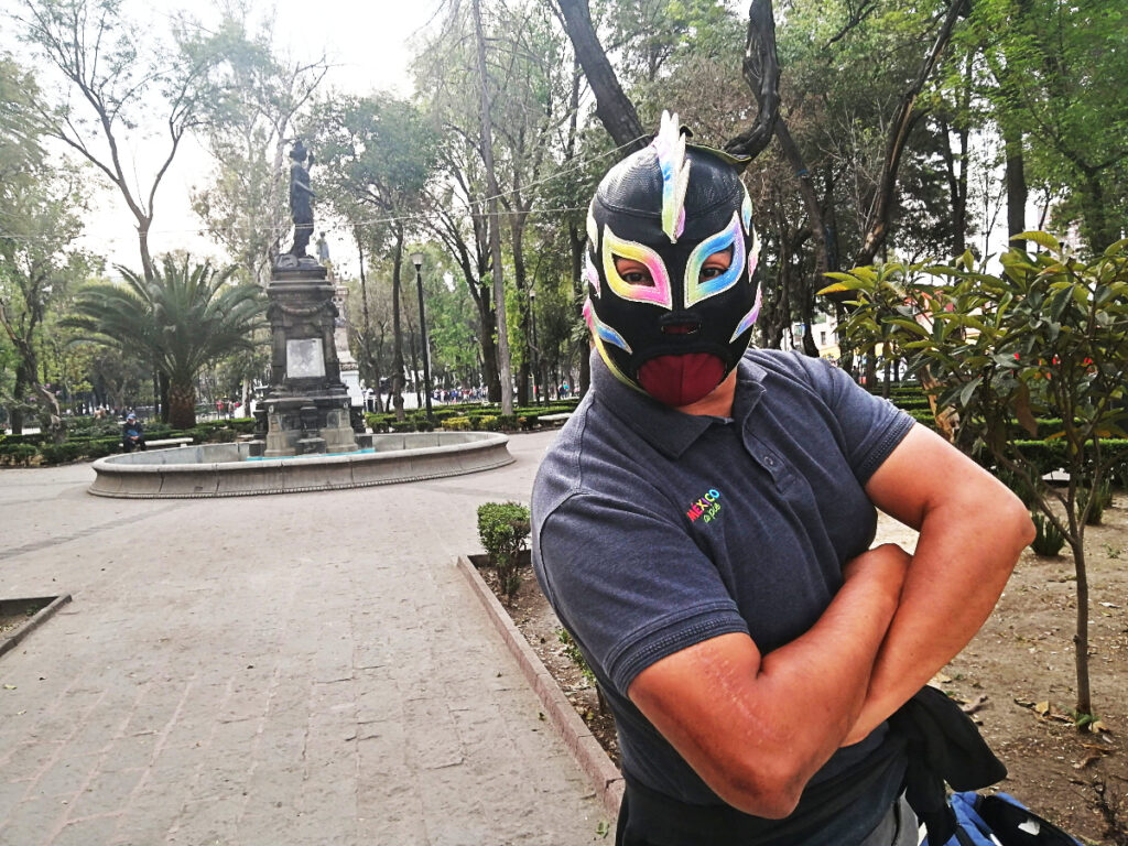 A former wrestler of Mexico City lucha libre poses wearing a black luchador mask highlighted with rainbow accents and a mohawk. His arms are folded in front of him.