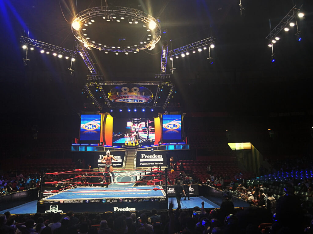 A luchador stands on the middle rope while igniting the crowd before lucha libre in Mexico City. Behind are large television screens to show the wrestling match. Above are track lighting including a large ring of light to light up the stage.