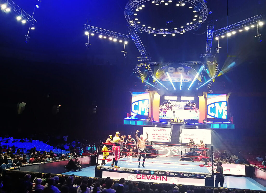 Several Mexican wrestlers take to the stage during lucha libre tour in Mexico City. Behind them are large TV screens to broadcast the wrestling match and lights to illuminate the stage.
