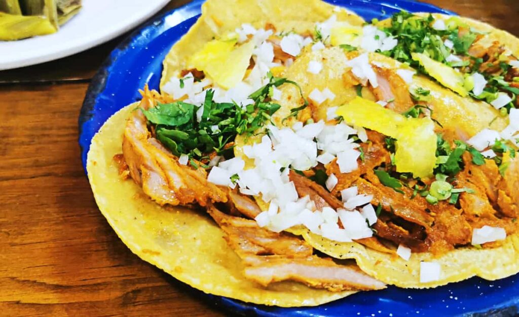 Four pastor tacos are layered on a round blue plate in Mexico City. They are topped with raw onion, cilantro, and pineapple.