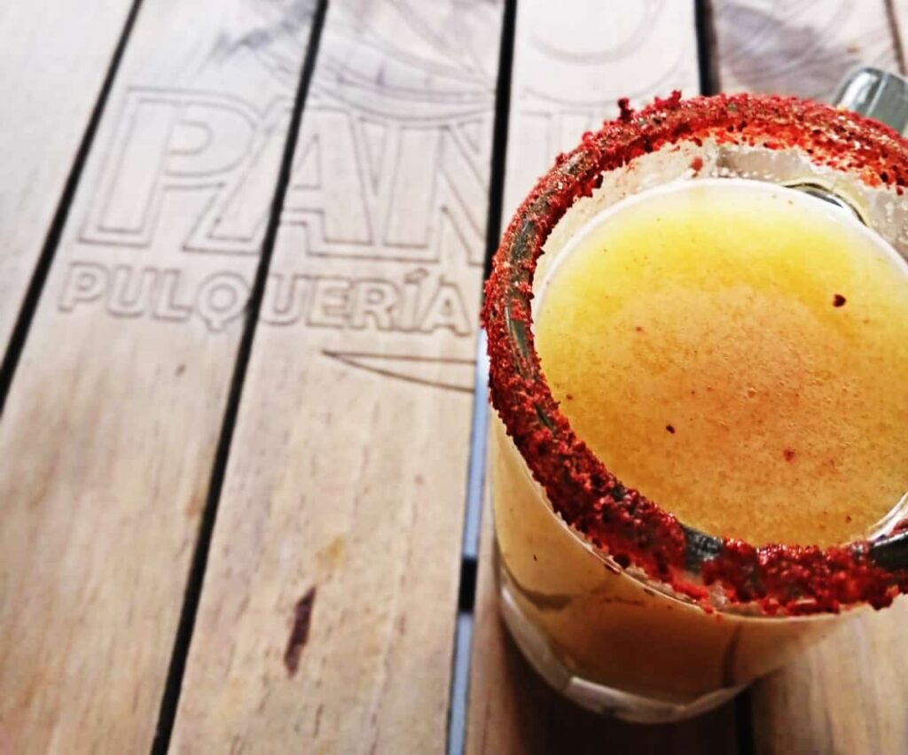 A yellow glass of pulque sits on a wooden table that is carved with the word Pulqueria. The pulque glass is rimmed with spicy chile salt.