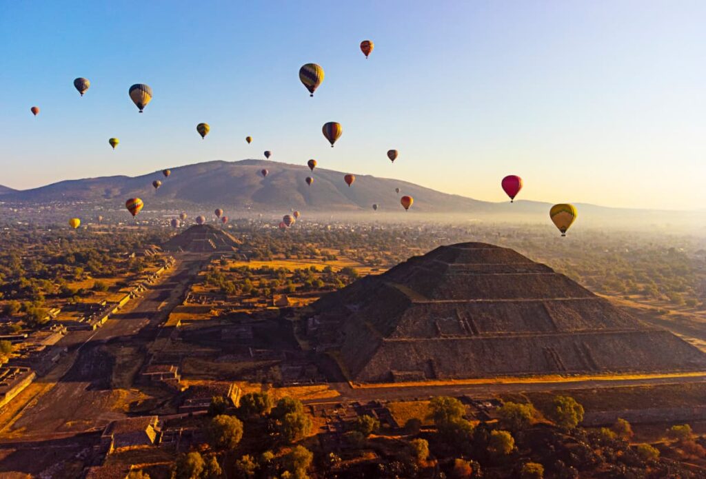 From Mexico City, hot air balloons ride over Teotihuacan in the early morning light. In the foreground is the Pyramid of the Sun with the Pyramid of the Moon in the background.