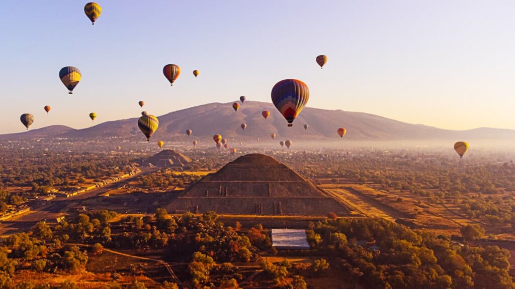 From Mexico City, hot air balloons fly over Teotihuacan pyramids. You can prominently see the Pyramid of the Sun with the Pyramid of the Moon in the background.