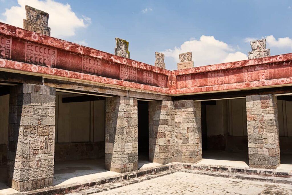 Inside the Place of Quetzalpapalotl at Teotihuacan features several rectangular carved stone columns. The red border above them is decorated with various symbols and topped with several stone decorations.