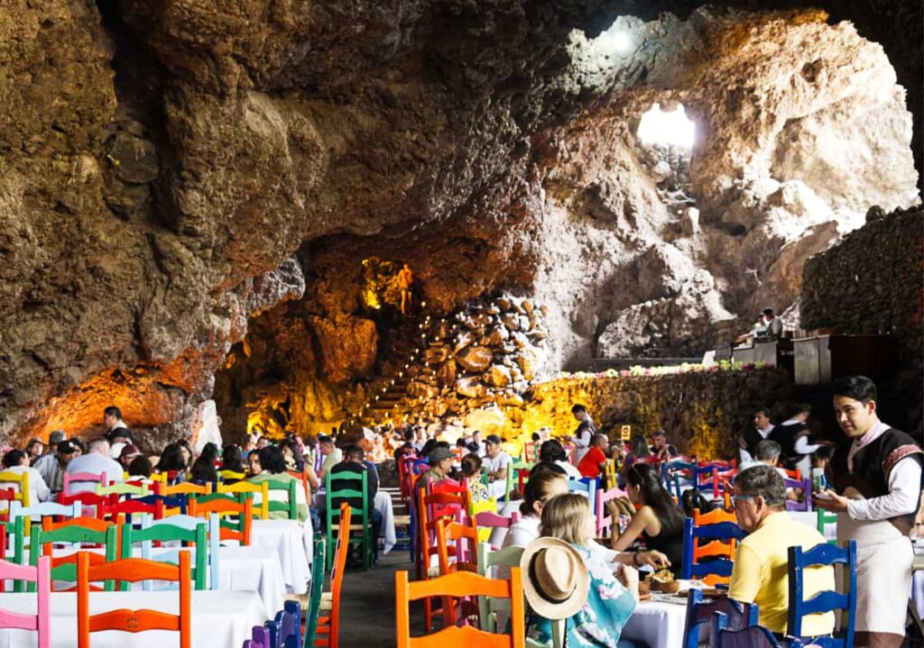 A waiter takes the order from a table of diners inside the cave restaurant at Teotihuacan Mexico. behind them are large stone formations that make up the cave, including a set of stairs where diners place a lit candle after their meal.