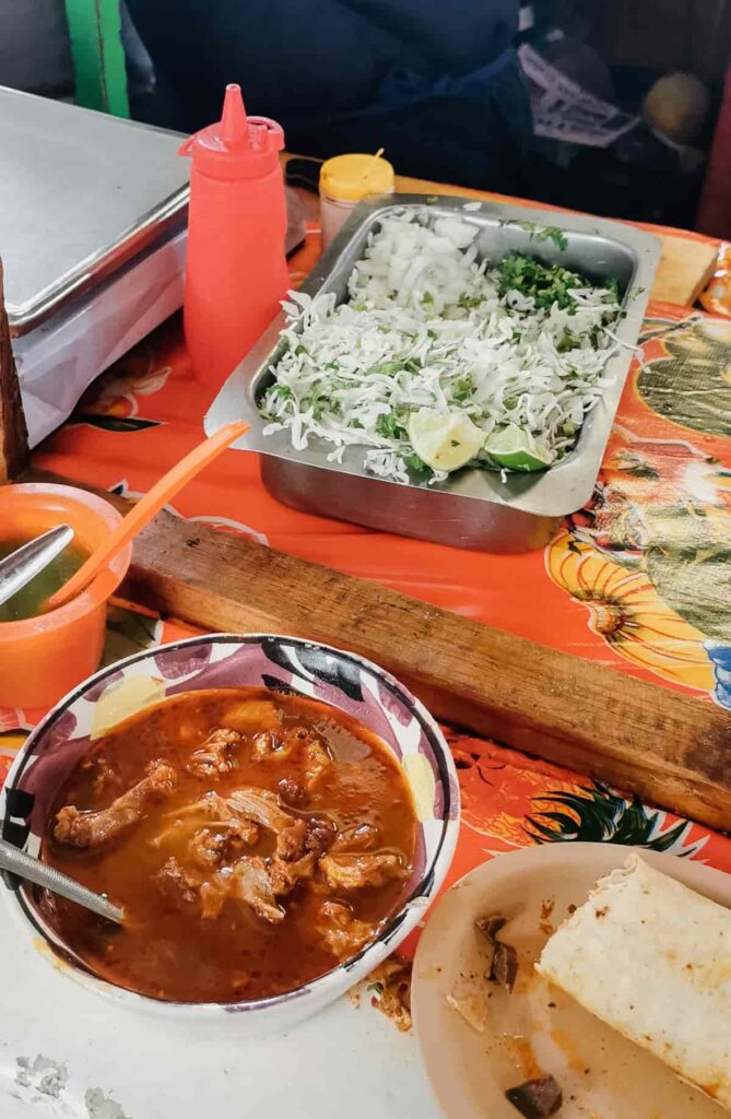 A barbacoa taco sits on a plate next to a bowl of consome during a taco tour in Mexico City. Behind is a container of shredded cabbage and cilantro.