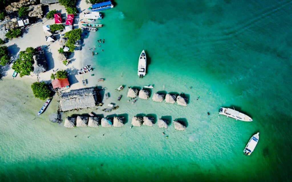 An overhead drone shot during a Cartagena boat tour shows several boats anchored in turquoise water off the coast of Isla Cholon. The edge of the island can be seen in the top left while in the water there are small huts with natural palapa roofs.
