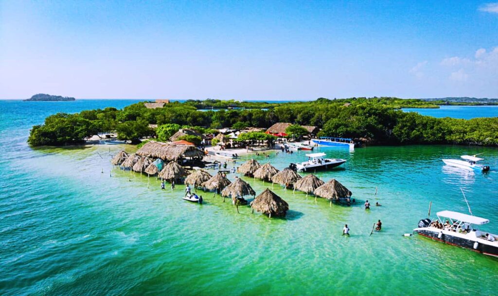 An ariel view from a Cartagena boat party show several small palapa huts in the ocean, tree tops on the edge of Isla Cholon, and several boats in the turquoise water.