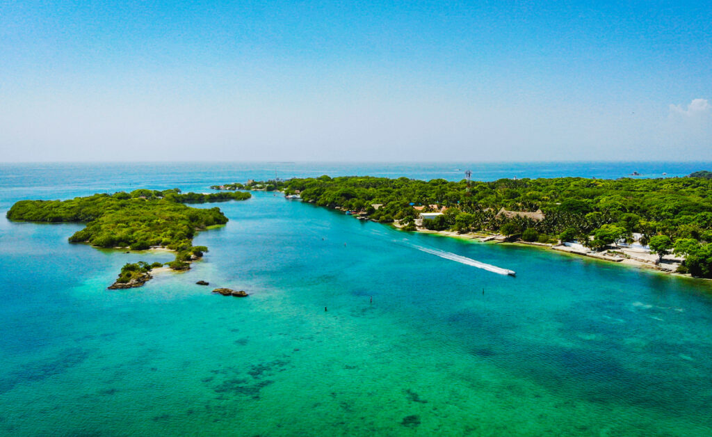 An ariel view of a speedboat passing through the Rosario Islands on a Cartagena island tour. The island behind the boat is filled with bright green treetops and a few houses. The other smaller island is full of trees.