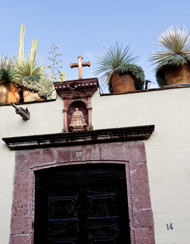 A stone framed doorway seen while walking in San Miguel de Allende. Above the door is a small stone figure with a stone cross lit by the sun. On the rooftop next to the cross are several potted plants.