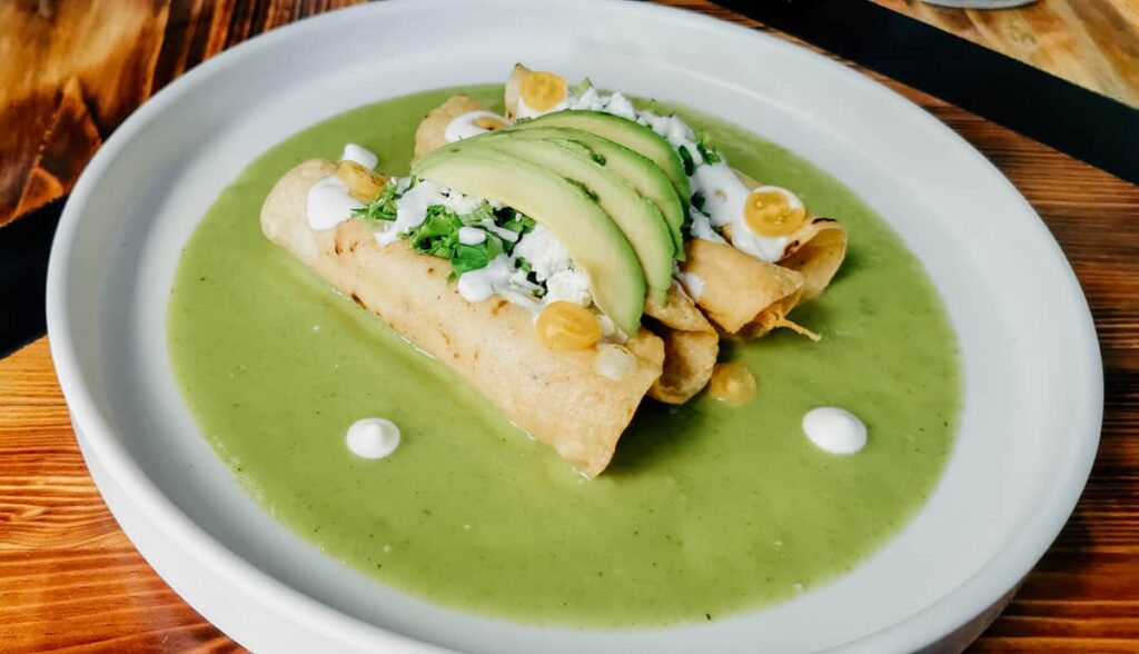 A plate of rolled and fried tacos dorados sit on top of a bright green avocado sauce during a taco tour in Mexico City. They are topped with fresh cheese, crema, and avocado.