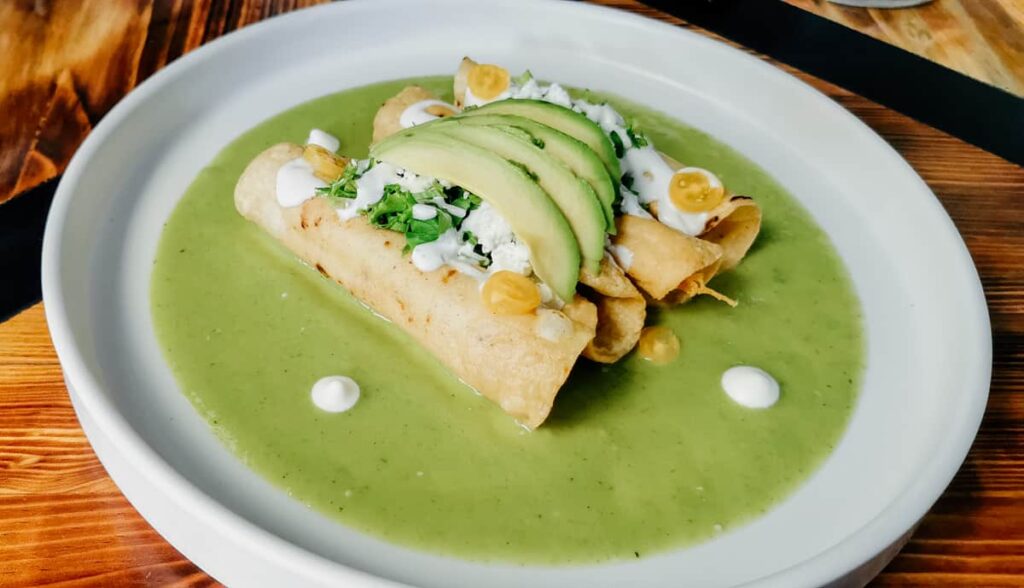 A plate of rolled and fried tacos dorados sit on top of a bright green avocado sauce San Miguel de Allende food tour. They are topped with fresh cheese, crema, and avocado.