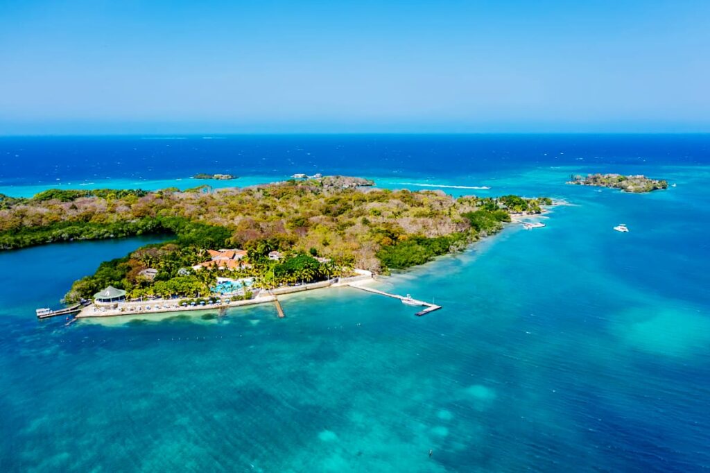 A ariel view of Isla Grande on a Cartagena island tour shows a large island with lots of trees, several houses, pool, and docks. The island is surrounding by vibrant blue and turquoise water.