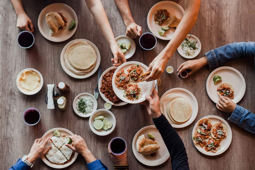 An overhead view of a Mexico City cooking class with several plates of food, tacos, limes, and salsa. Several hands reach out for a plate of al pastor tacos.