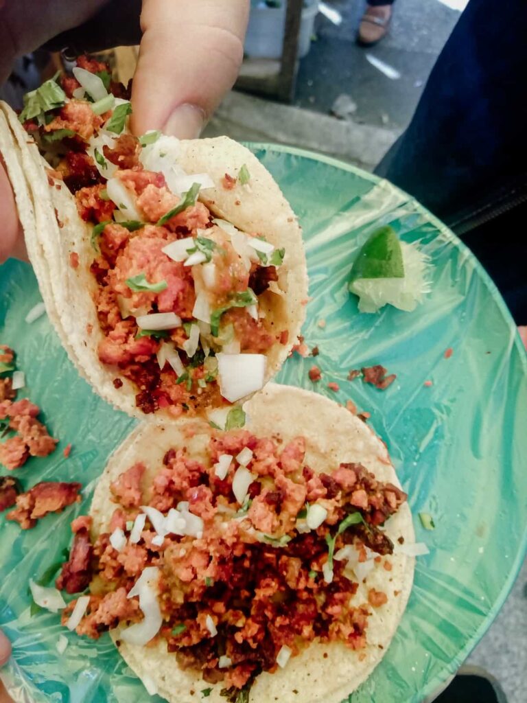 Eating tacos during a Mexico City taco tour, a hand picks up one taco full of meat and topped with onion and cilantro while the other sits on the plate below.