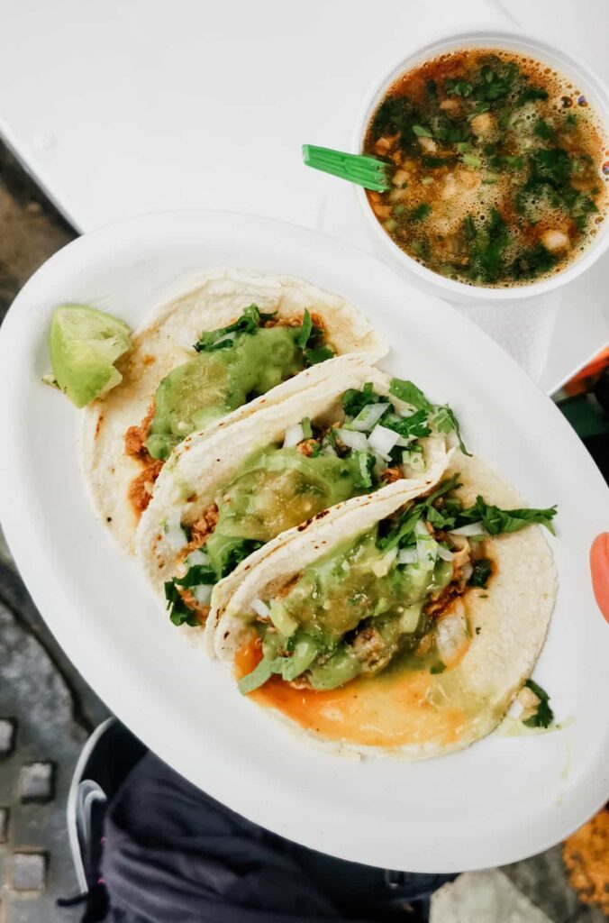 A plate of three barbacoa tacos line a plate next to a cup of consome on a taco tour, Mexico City style.