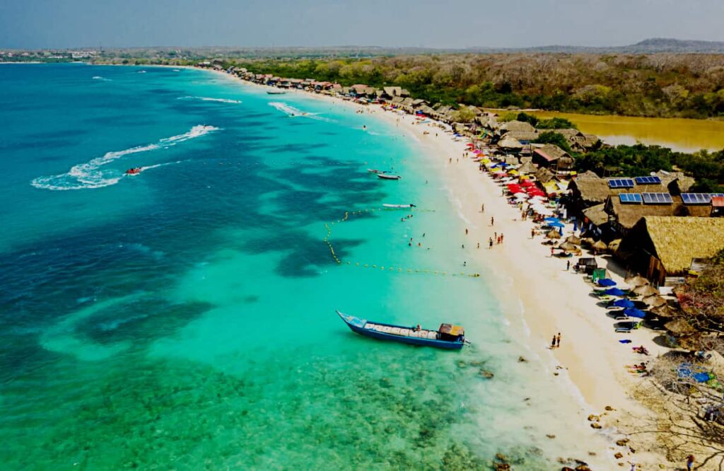 An ariel view of Playa Blanca Baru, one of the stops on the Cartagena island tours shows bright turquoise water and a long stretch of beach. The beach is filled with people, chairs and umbrellas, and buildings with palapa roofs. A boat is anchored in the water in the foreground.