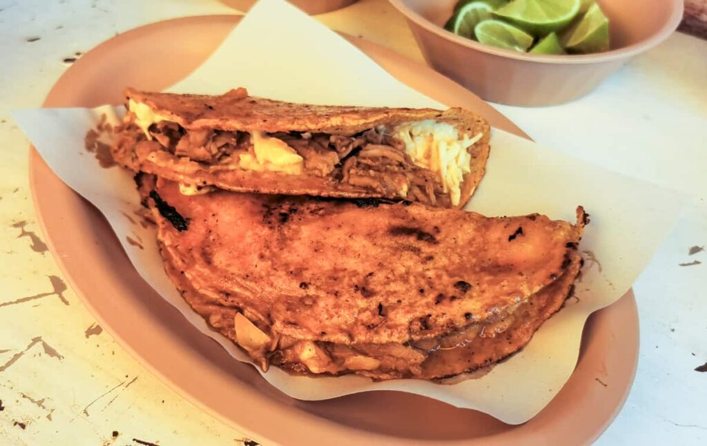 Juicy, greasy quesabirria tacos on a plate during a taco tour in Mexico City. In the background is a bowl of cut limes.