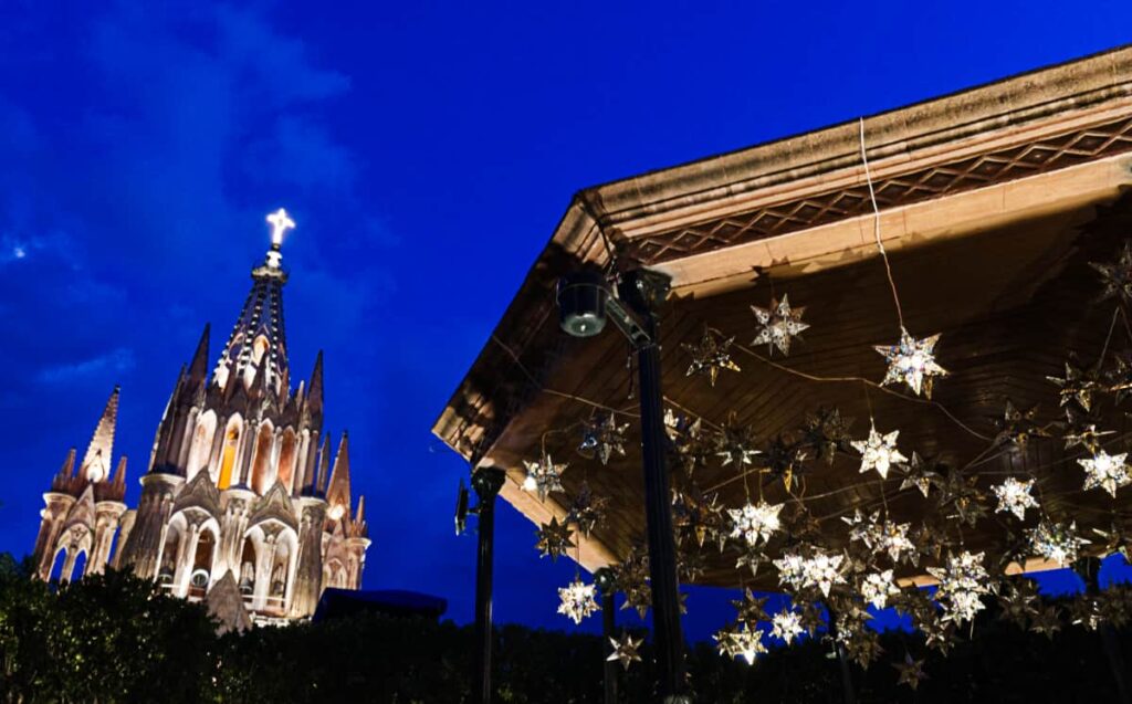 The gazebo in the main park of San Miguel de Allende is lit with hanging star decorations. In the background, the main church is lit up at night.